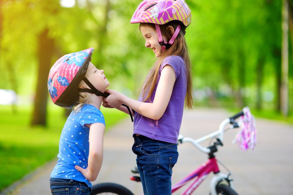 Two young smiling girls wearing helmets stand on a tree-lined path with a bicycle in the background. The older girl, in a purple shirt and jeans, is adjusting the helmet of the younger girl, who is wearing a blue shirt with white polka dots. 