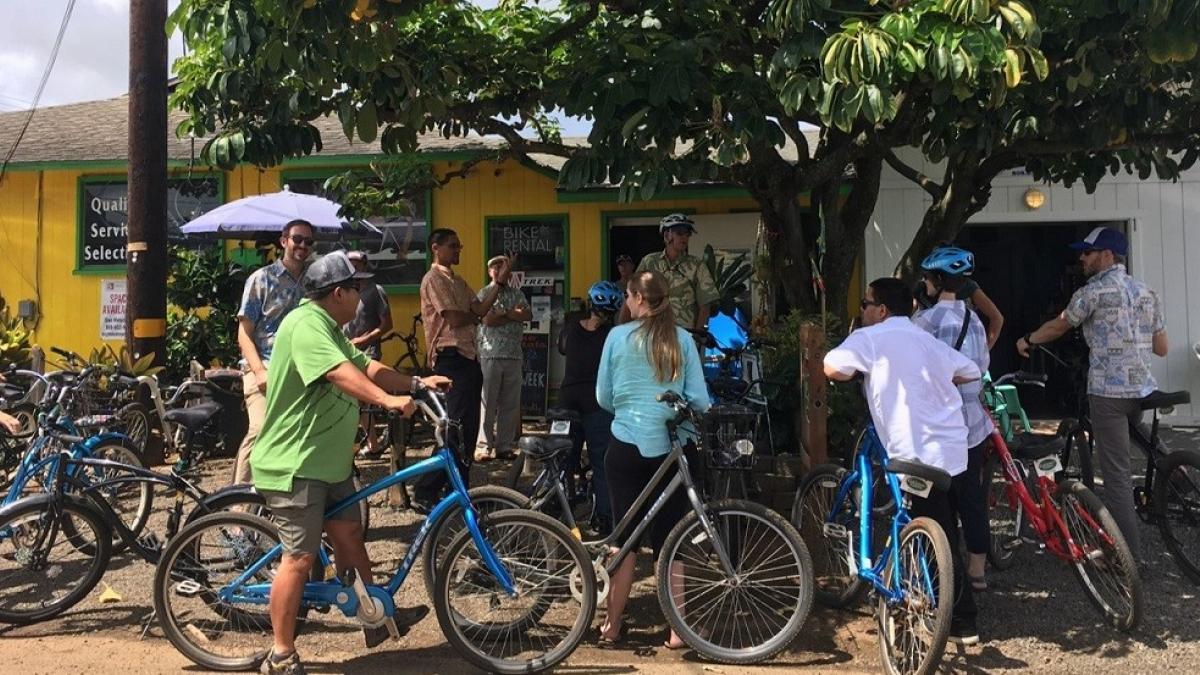 People at a bicycle rental shop outdoors, with a range of bicycles in the foreground and a yellow rental building in the background.